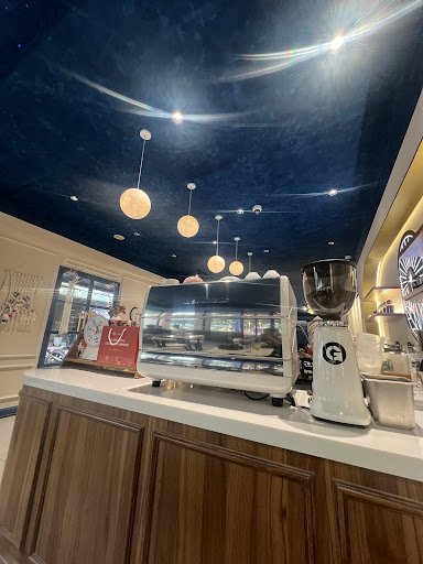 The image shows a modern coffee shop interior featuring wooden countertops and ceiling decor that mimics the night sky with stars and hanging spherica