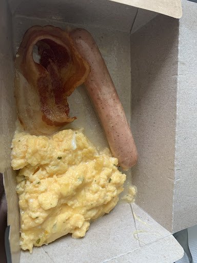 The image shows a takeaway food item consisting of scrambled eggs and two sausages in a cardboard container.