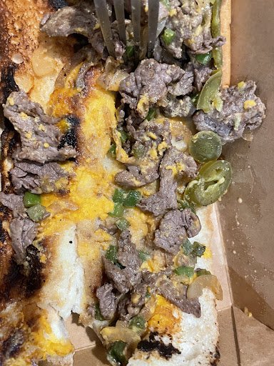 A close-up of a dish consisting of grilled steak topped with melted cheese and green chilies on what appears to be bread or flatbread, accompanied by 