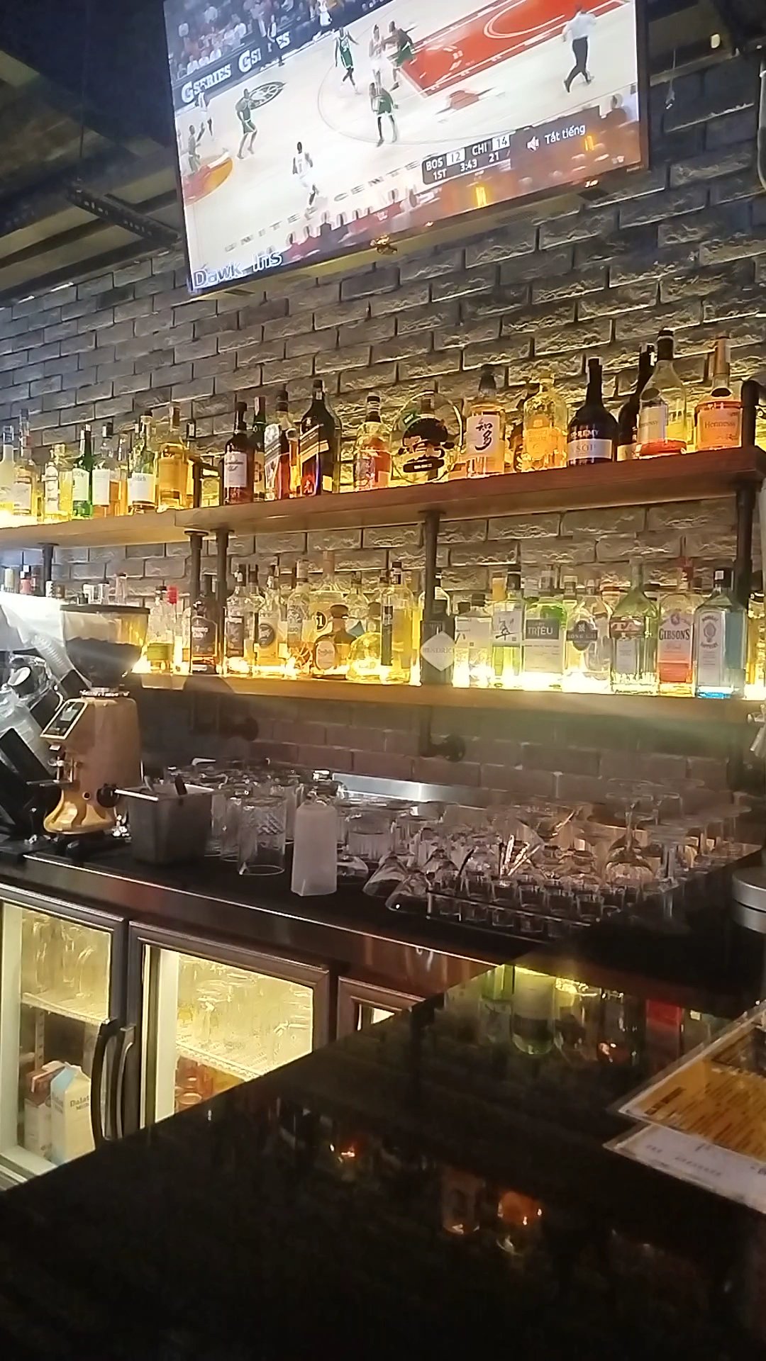 A bar interior featuring multiple shelves lined with various liquor bottles against a brick wall, a large television displaying a basketball game, a c