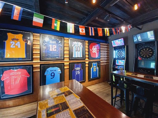 The image shows an interior of a sports bar or pub featuring framed jerseys on the wall and arcade games in the background.
