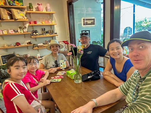 A group of people is gathered around a wooden table in what appears to be an indoor cafe or restaurant setting with shelves displaying various items a