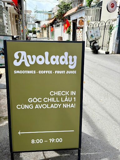 A green signboard for 'Avolady', a smoothies and coffee shop located on an urban street with buildings, flags, and scooters in the background.