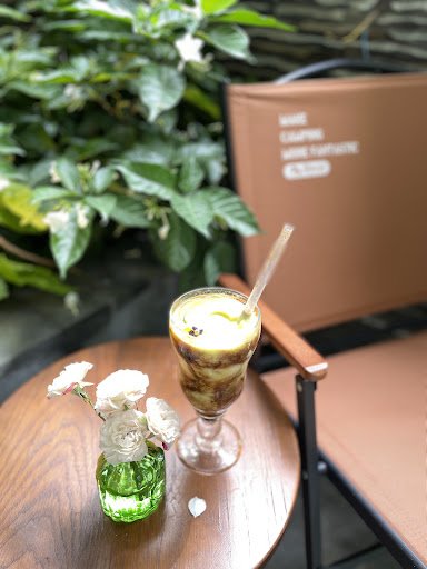 A glass of layered coffee beverage sits on a wooden table beside a small vase containing white flowers and green liquid. In the background, there is a
