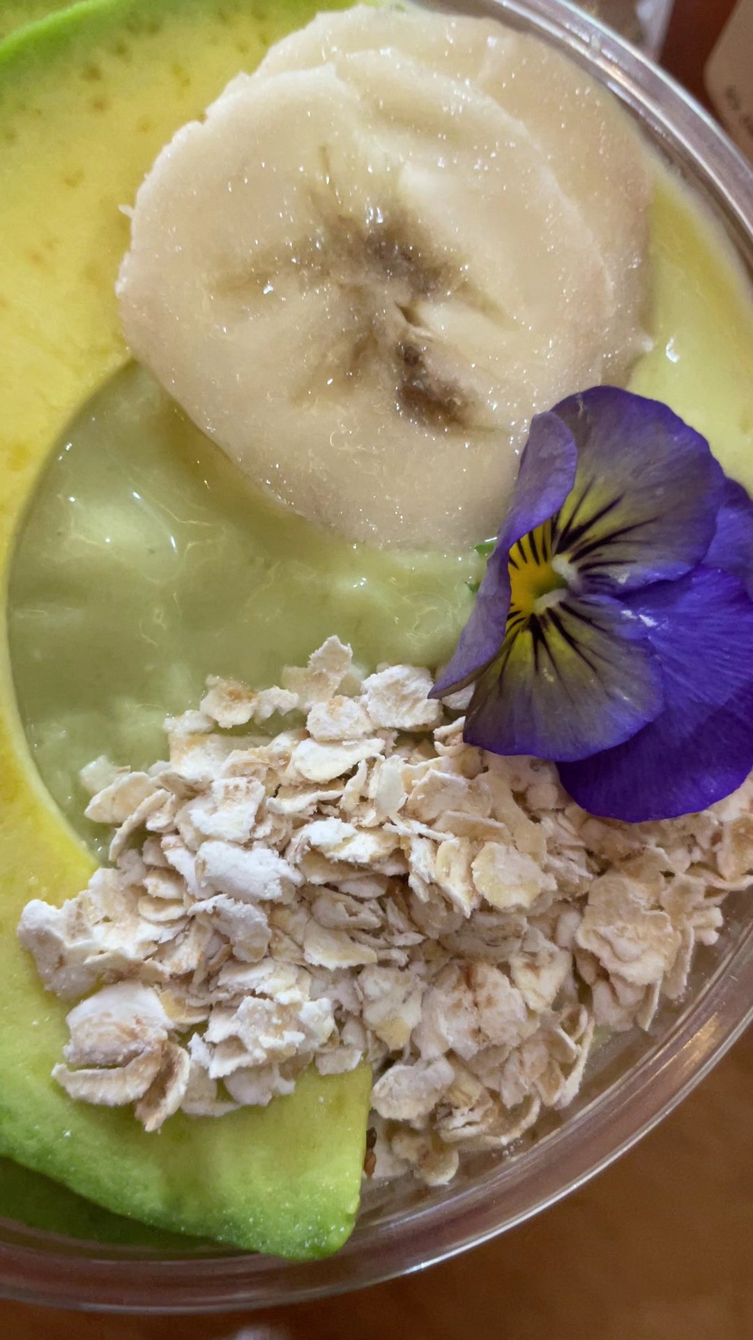 A close-up of a bowl containing oat flakes on top of avocado slices and banana pieces, garnished with a purple pansy flower.