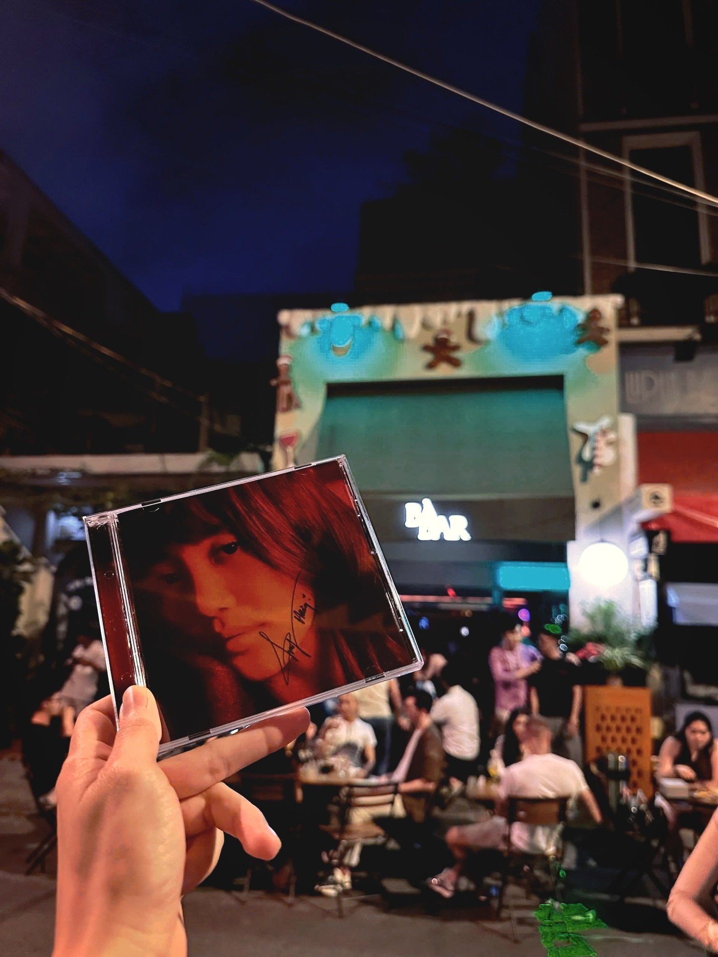 A person is holding up a CD case in front of an outdoor venue during nighttime with people sitting at tables and engaging in conversations or dining.