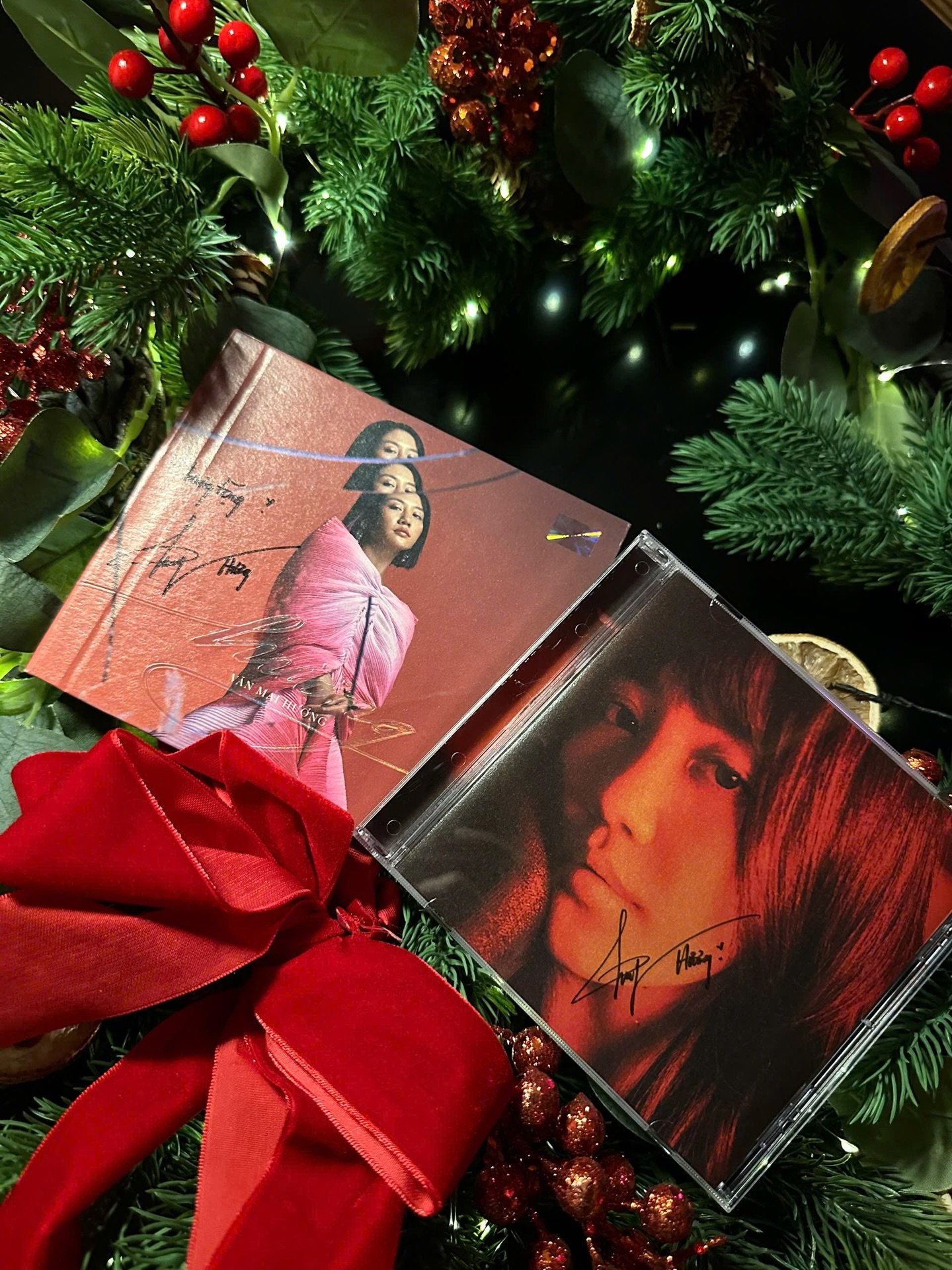 The image shows a close-up of two CD cases placed on top of each other with Christmas decorations in the background including red berries, holly leave