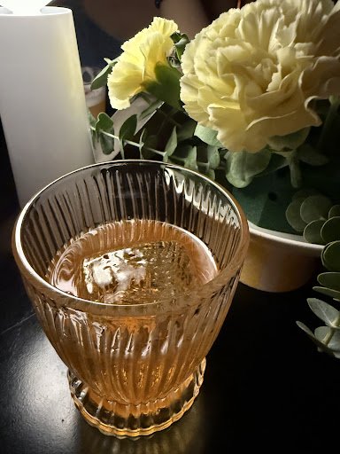 A close-up of a glass containing an amber-colored beverage on a dark surface, with ice cubes visible inside the glass. In the background, there is a v