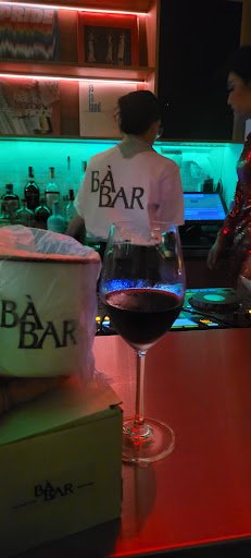 A person wearing a white 'BAR BAR' shirt is standing behind the bar counter in an establishment with vibrant lighting and various bottles on display.