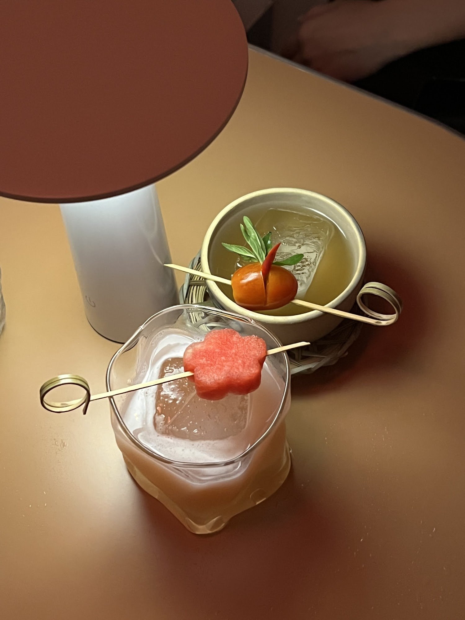 The image shows a close-up of two drinks on a table in what appears to be an upscale venue with dim lighting and warm tones, suggesting it could be ev