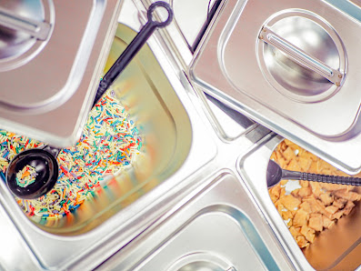 The image depicts a close-up view of a dessert station in a venue, featuring multiple metal containers. One container holds colorful sprinkles, anothe