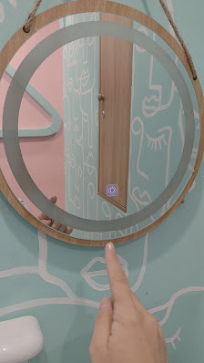 A hand is pointing at a sticker on an oval-shaped mirror hanging above a toilet in a bathroom with pink and white walls.