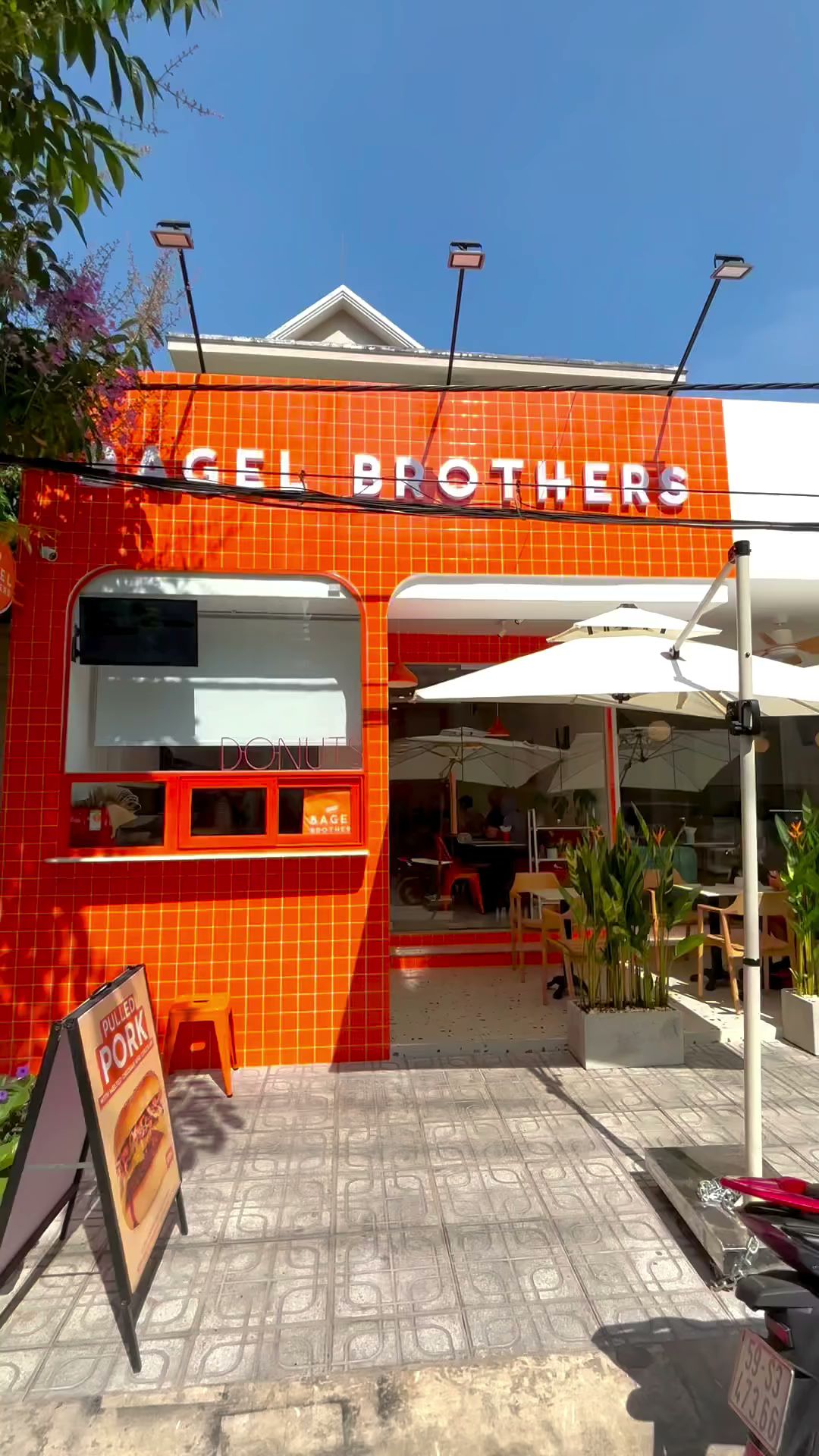 The image shows an exterior view of a brightly colored building named 'Bagel Brothers' with orange tiled walls and white trimmings. The establishment 