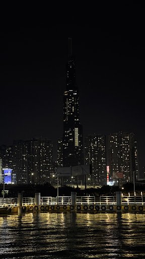 The image captures a nighttime cityscape featuring an illuminated skyscraper and surrounding high-rise buildings reflected in the water of what appear