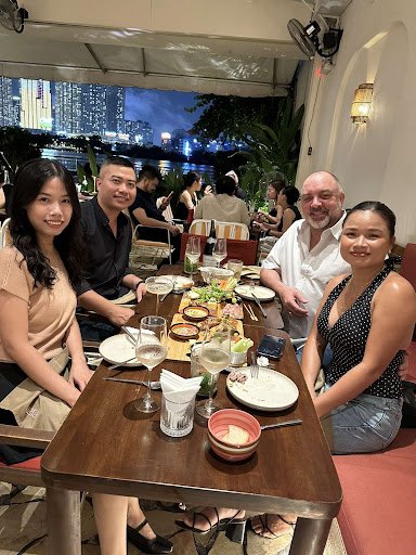 A group of people is seated at a dining table in an outdoor restaurant setting during the evening with city lights visible in the background.