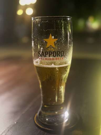 A close-up shot of a glass filled with Sapporo Premium Beer on a wooden surface, likely taken in an indoor setting given the blurred lights and bokeh