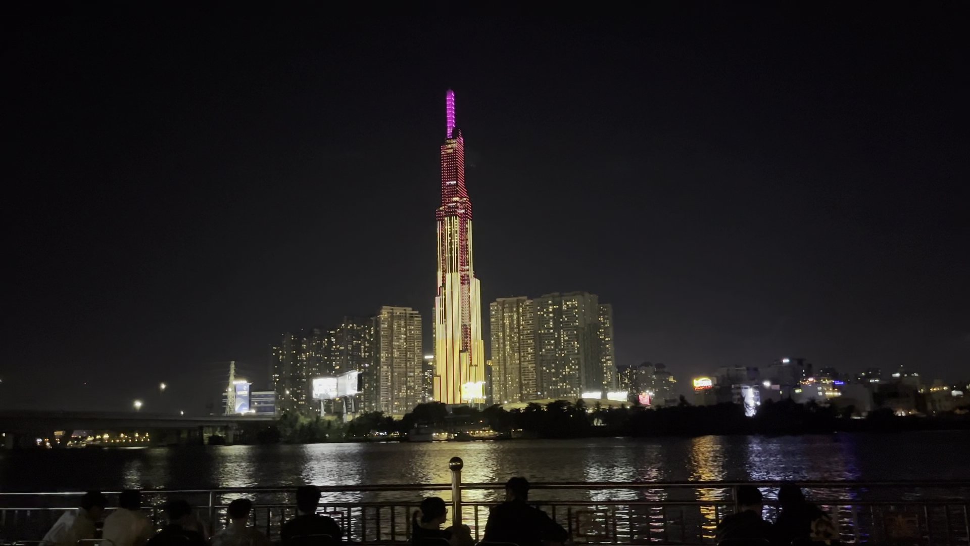 The image captures a nighttime cityscape featuring an illuminated skyscraper as the focal point. The building is adorned with vibrant lights, includin