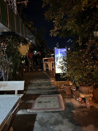 The image depicts an outdoor seating area of a restaurant or bar at night. There are several potted plants and wooden stools visible, with dim lightin
