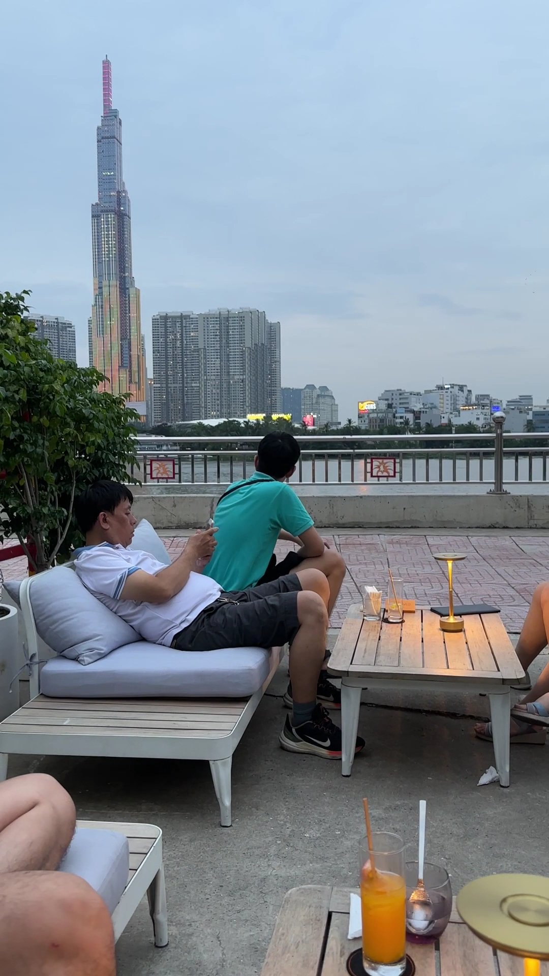 The image captures a group of people relaxing on an outdoor terrace overlooking the Saigon River and prominent skyscrapers in Thao Dien, Saigon.
