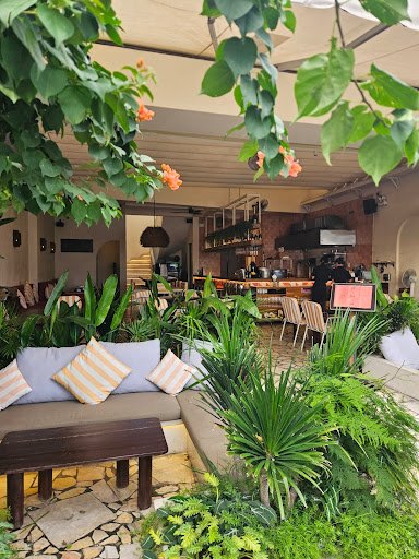 The image shows an interior view of a cozy café or restaurant in Thao Dien, Saigon. The space is adorned with lush green plants and vibrant flowers th