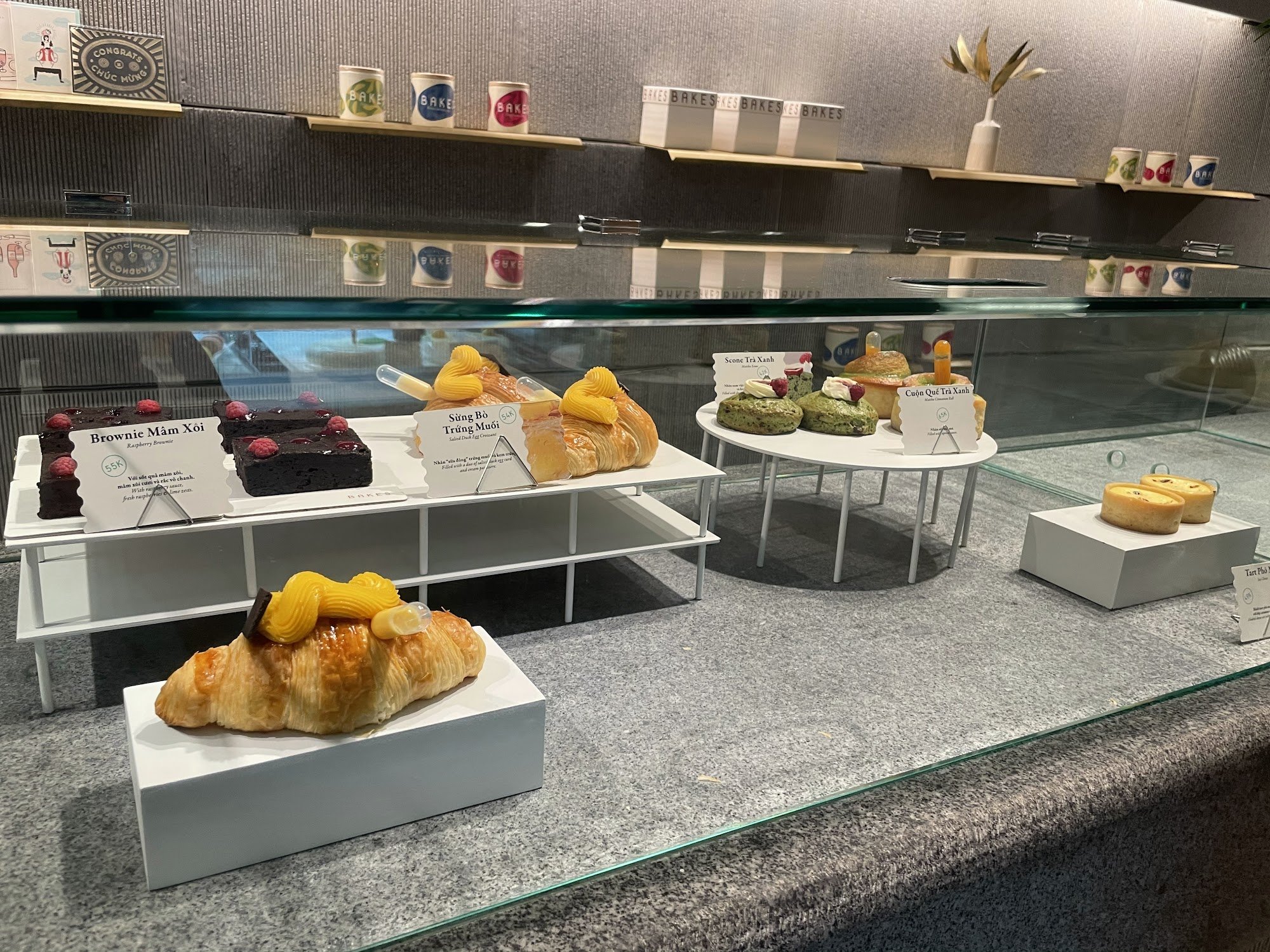 The image depicts the interior of a bakery or café in Thao Dien, Saigon, featuring a glass display case with various pastries and cakes. Visible items