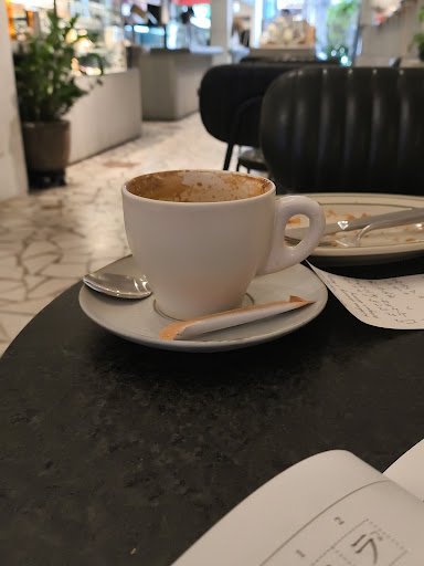 A close-up of a coffee cup on a saucer in what appears to be an indoor cafe setting with modern decor, including dark leather chairs and marble tables