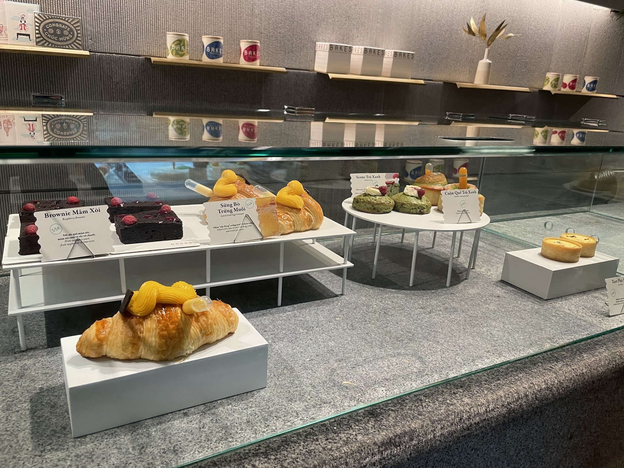 A glass display case in a bakery/cafeteria showcasing various pastries and cakes. Items include Brownie Mâm Xôi (raspberry brownies), Súng Bồ Trứng Mu