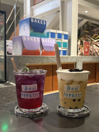 Two beverages are displayed on a counter in front of the 'BAKES' branding boxes. The drinks have labels with French text that translates to 'Bon Appet
