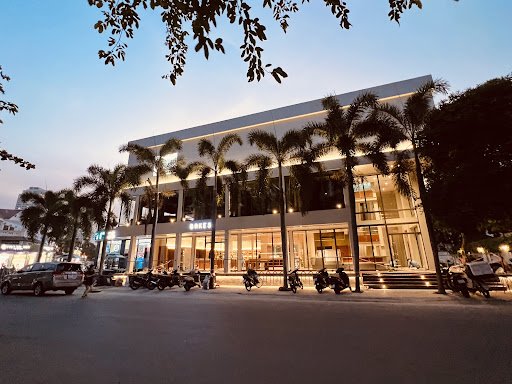The image depicts a modern building at dusk or early evening in Thao Dien, Saigon. The structure has large glass windows and is well-lit from within, 