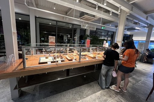 The image shows an interior view of a bakery or café where customers are browsing pastries displayed in a glass case.