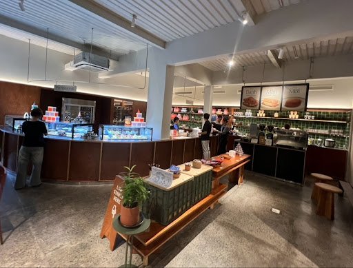The image depicts an interior view of a modern coffee shop or bakery store. The space is well-lit with overhead lighting and features wooden counters,