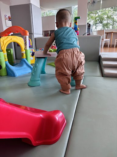 A young child is standing on a play mat in what appears to be a living room or family area of an apartment, with various toys and furniture items visi