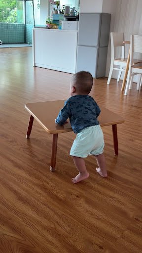A young child is standing on a wooden floor and leaning against a small table in what appears to be a modern kitchen or dining area with light-colored