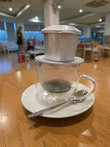 The image shows a clear glass cup containing what appears to be coffee or tea placed on top of another transparent container with a metal lid, possibl