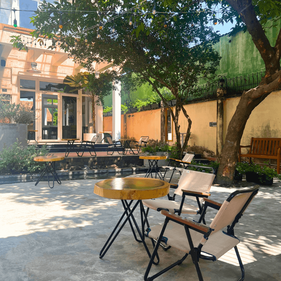 Outdoor courtyard of a venue in Thao Dien, Saigon. Features concrete patio, wooden tables with black metal legs, beige cushioned chairs, string lights