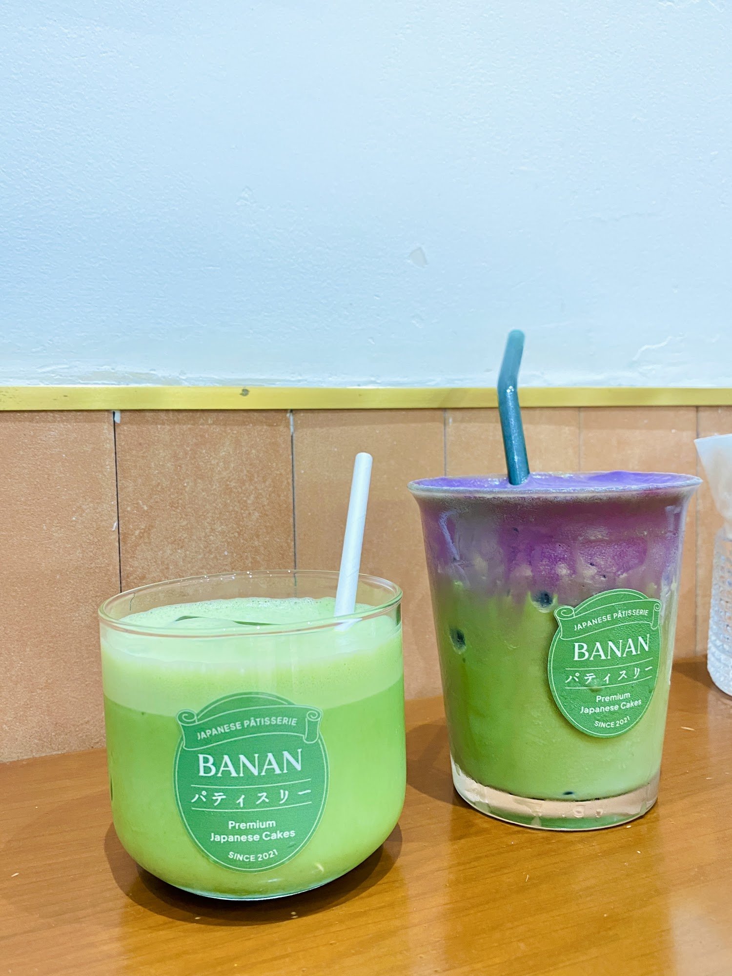 Two beverages from a Japanese patisserie are displayed on a wooden surface against a light blue wall with beige tiles.