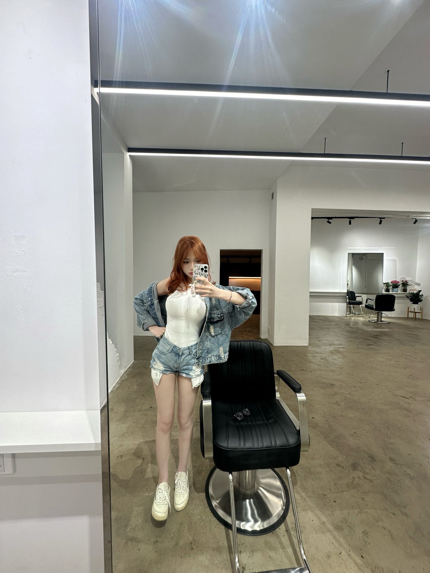 A woman is taking a selfie in front of a large mirror inside what appears to be a modern hair salon or barbershop.