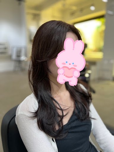 A woman is seated in what appears to be a modern indoor setting, possibly a cafe or waiting area with blurred background elements suggesting other pat