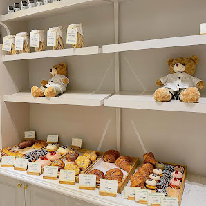 The image shows a well-lit interior display of desserts and pastries on shelves with teddy bears dressed in white shirts, adding to the aesthetic appe