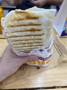 A hand is holding a folded wrap or flatbread that appears to be toasted and has grill marks on it. The food item is placed in a paper container with r