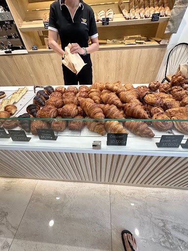 The image shows a bakery counter displaying an array of baked goods such as croissants and other pastries on a glass display case, with price tags vis