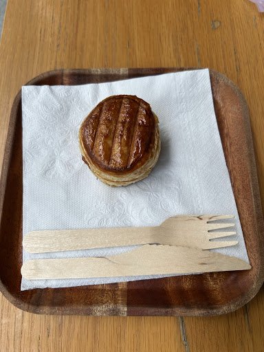 A single pastry topped with a caramelized topping sits on a white paper napkin within a wooden tray. Beside the pastry, there is a set of wooden utens