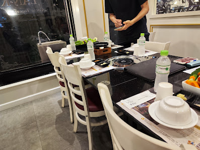 A restaurant interior in Thao Dien, Saigon, featuring a dining table set with white chairs, plates, bowls, water bottles, and a bowl of fruit. A perso