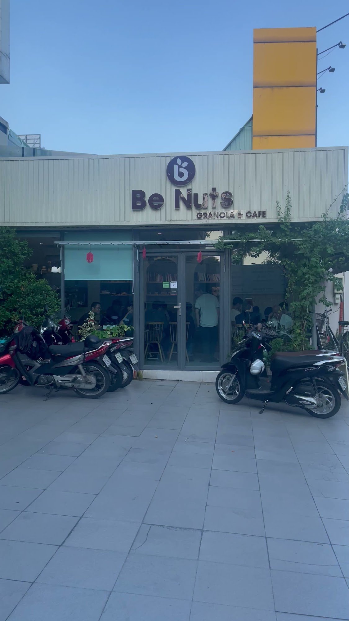 The image shows an exterior view of a cafe named 'Be Nuts Granola & Cafe'. The establishment has a modern facade with large windows and glass doors, t