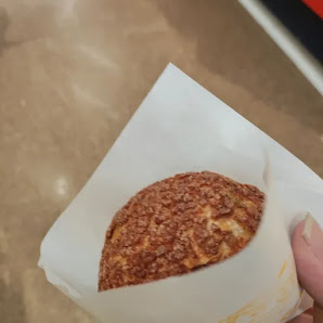 A hand is holding a pastry wrapped in paper that appears to be freshly baked and topped with what looks like crushed nuts or seeds.