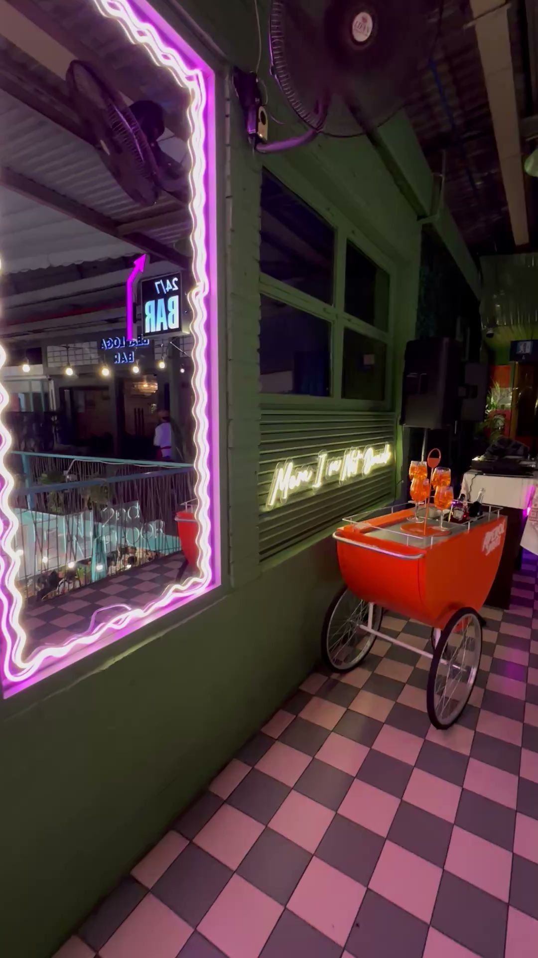 The image shows an interior view of a bar or restaurant at night. Neon lights outline the windows and walls in vibrant colors, creating a lively atmos