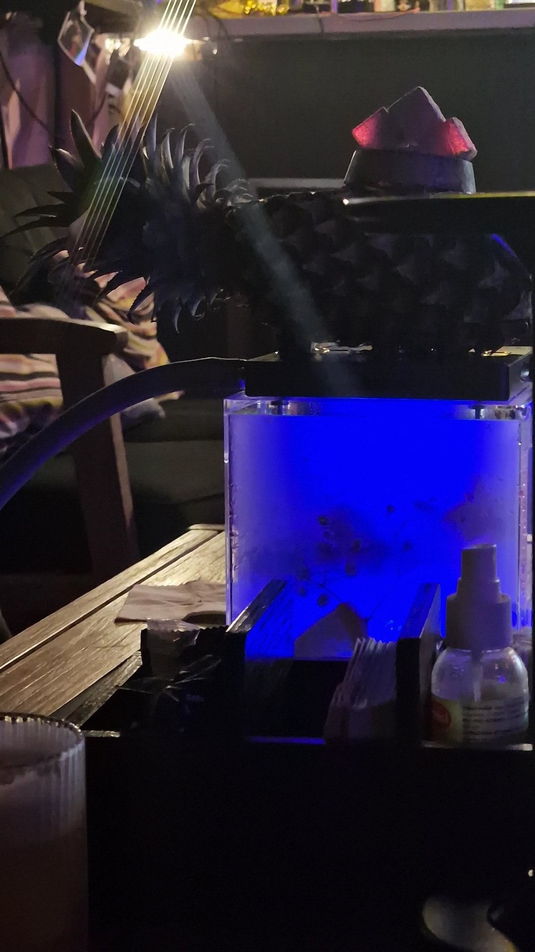 A dimly lit interior scene featuring a blue-lit aquarium on a table where someone is playing an electric guitar with a hat placed atop it.