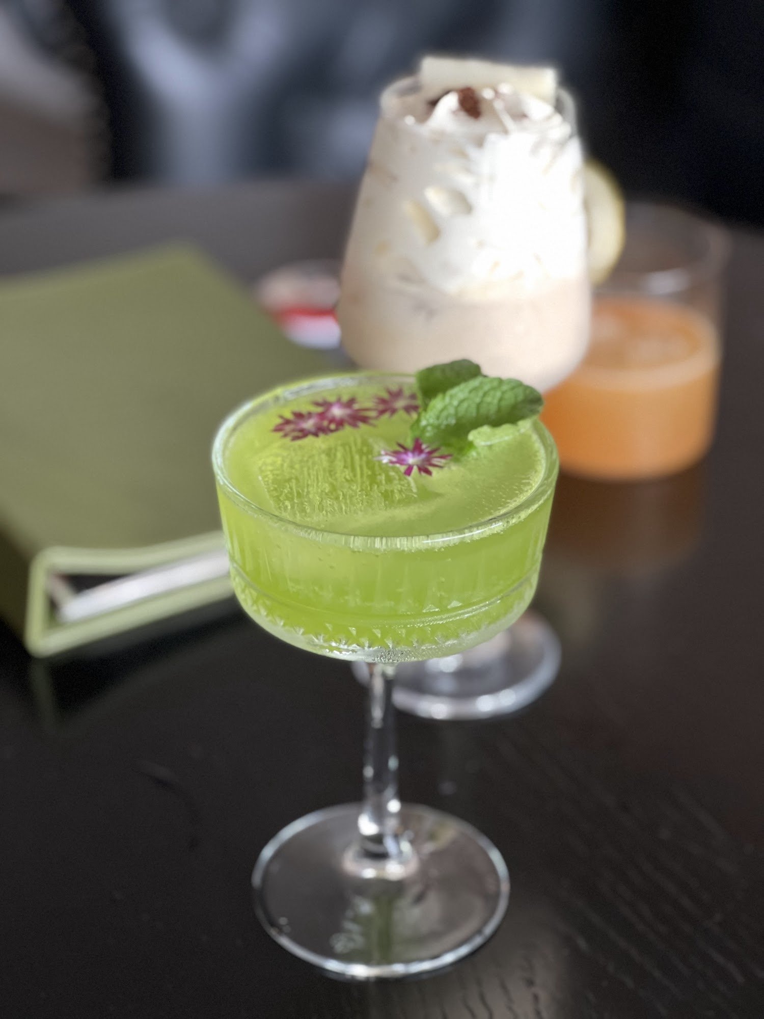 A close-up of a bright green cocktail garnished with mint and edible flowers in a clear glass on a dark wooden table, accompanied by other drinks out