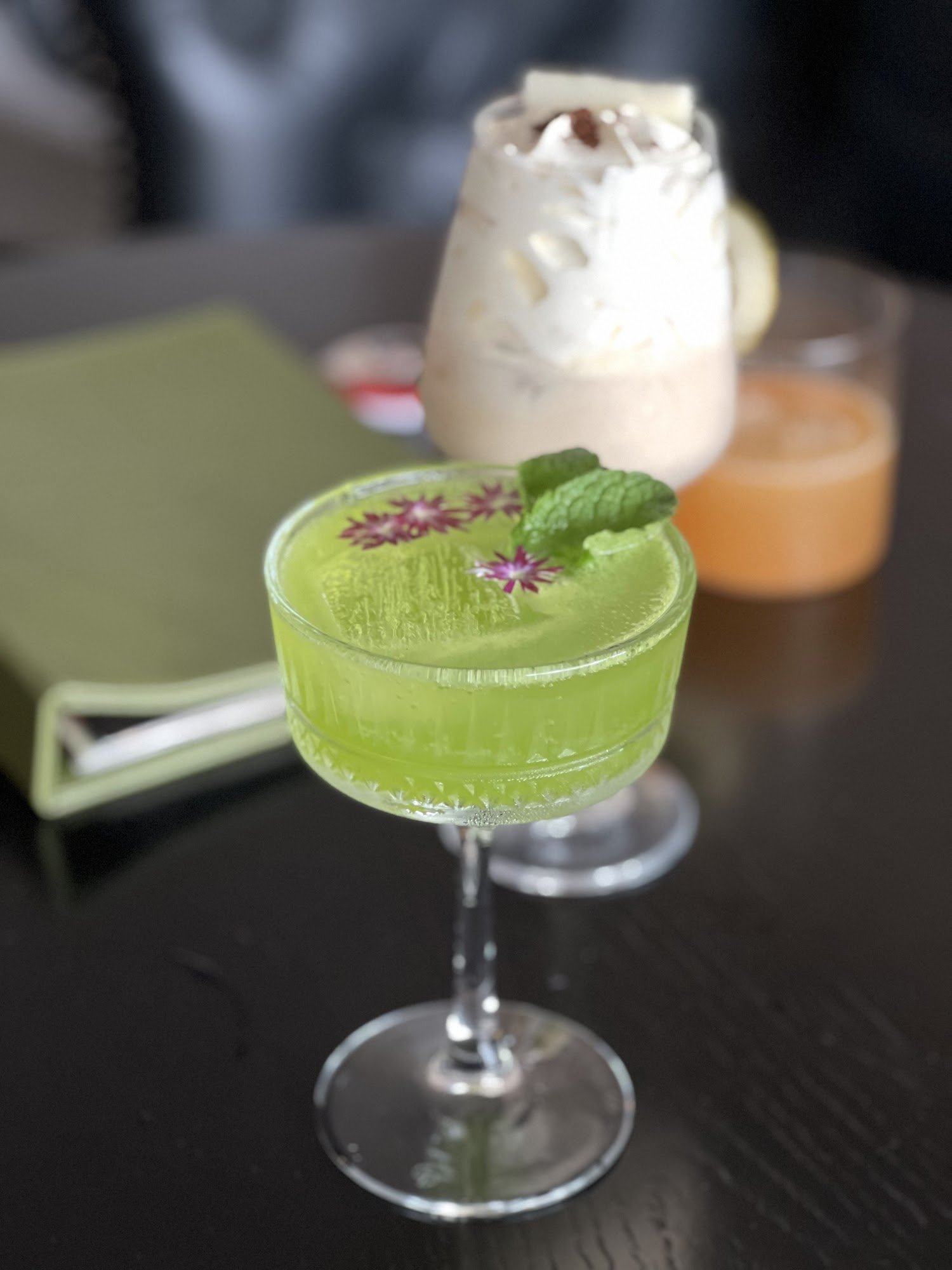 A close-up shot of a vibrant green cocktail garnished with mint leaves and edible flowers in a clear glass on a dark table, accompanied by other drink
