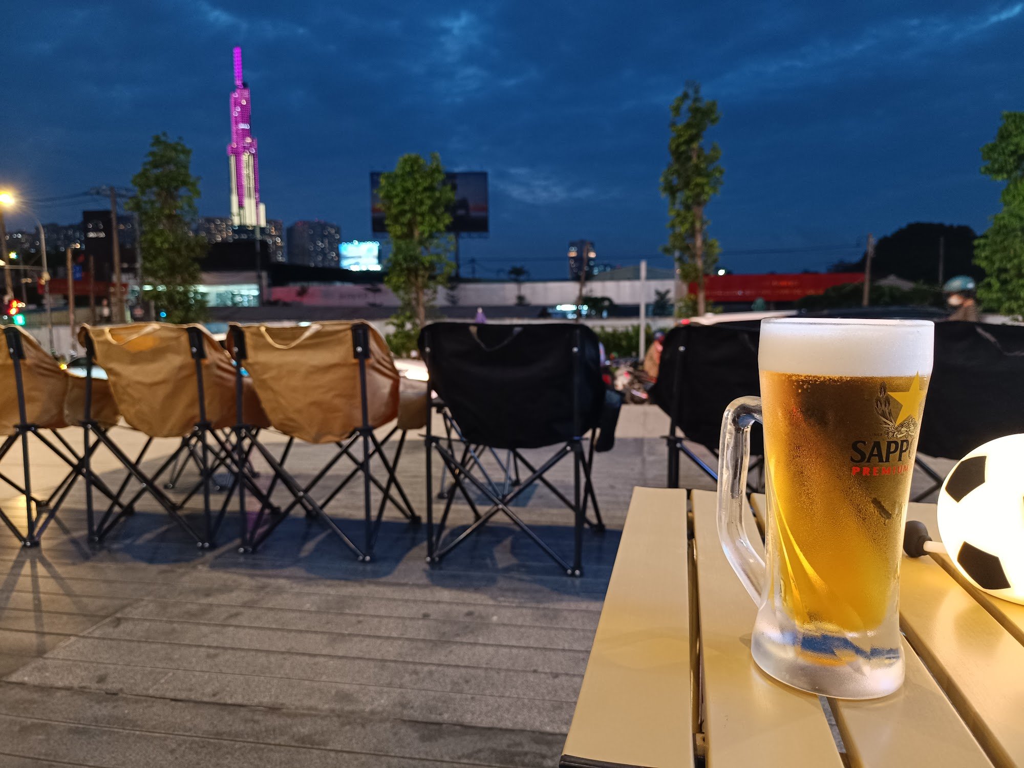 The image captures an outdoor seating area during twilight hours at Thao Dien in Saigon. The foreground shows a glass of Sapporo beer on a table, with